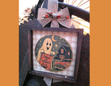 A framed Halloween wall hanging with a dark wood frame, burlap ribbon bow, and a central circular illustration of a ghost holding a 'Trick or Treat' sign in front of a haunted house and moon.