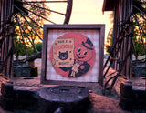 A square framed vintage-style Halloween sign with a round illustration reading 'HAVE A SPOOKTACULAR NIGHT!' featuring a black cat and a pumpkin character, displayed outdoors on a rustic metal structure at sunset.