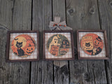 Three framed Halloween art prints displayed side-by-side on a rustic weathered wood background. The left print shows a black cat and jack-o'-lantern in front of an orange full moon.